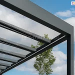 Zbliżenie na górny narożnik i konstrukcję dachu nowoczesnego zadaszenia tarasowego Spekta Glass, ukazane na tle błękitnego nieba z rozproszonymi białymi chmurami. Konstrukcja nośna została wykonana z solidnych profili aluminiowych w kolorze antracytowym o subtelnej, matowej teksturze, tworzących geometryczną ramę wspartą na pionowym słupie. Dach zadaszenia składa się z równolegle rozmieszczonych krokwi aluminiowych, na których zamontowano tafle w pełni przeziernego, bezpiecznego szkła. System szklanych paneli zapewnia maksymalny dostęp światła dziennego do przestrzeni pod zadaszeniem, jednocześnie chroniąc ją przed opadami. Pomiędzy profilami dachu widoczna jest korona zielonego drzewa, co podkreśla minimalistyczny design konstrukcji, która harmonijnie wtapia się w otoczenie. Całość prezentuje techniczny detal łączenia profili oraz wysoką jakość wykończenia elementów systemowych.