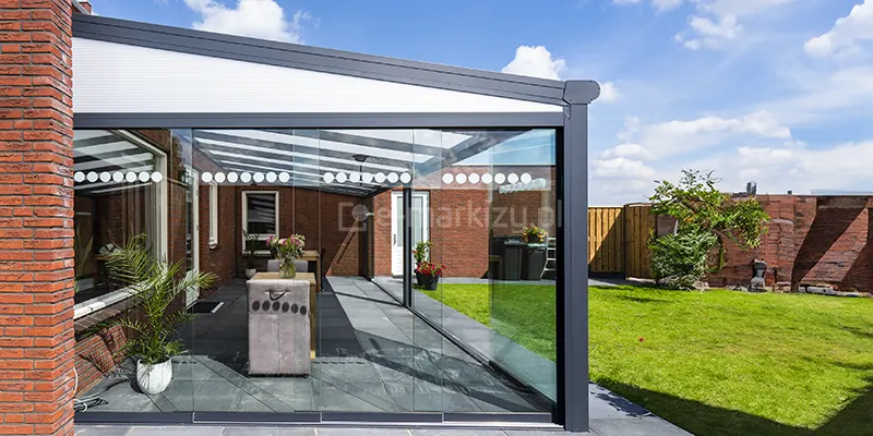Zdjęcie prezentuje boczną perspektywę nowoczesnego zadaszenia tarasowego zamontowanego przy budynku z czerwonej cegły. Konstrukcja w kolorze antracytowym posiada jednospadowy dach z jasnym wypełnieniem oraz system przesuwnych szyb bocznych, które tworzą zamknięty ogród zimowy. Na szklanych panelach umieszczono dekoracyjne rzędy białych kółek, pełniące funkcję ostrzegawczą i estetyczną. Wewnątrz tarasu, wyłożonego szarymi płytami, widoczne są meble ogrodowe oraz rośliny w donicach, a na zewnątrz rozciąga się zielony trawnik ogrodzony drewnianym płotem pod błękitnym niebem.
