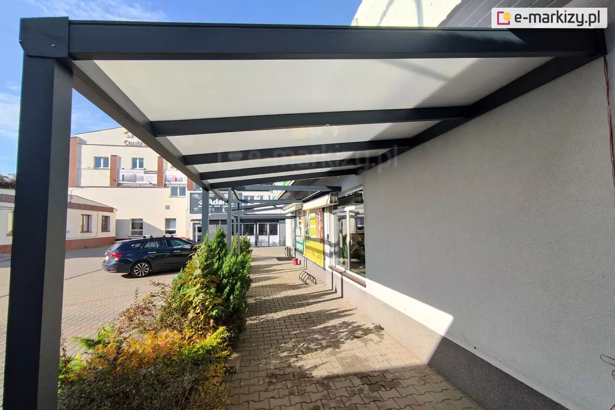 Zadaszenie z poliwęglanu Modes przymocowane do ściany, wypełnione poliwęglanem komorowym o mlecznej barwie osłania przejście wzdłuż ściany. Pod zadaszeniem kostka brukowa, do której przymocowano stojak na rowery. Przy krawędzi zadaszenia posadzone liczne rośliny zielone. W tle widoczny budynek z szerokim wejściem oraz zaparkowany w pobliżu samochód.