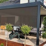 Zdjęcie przedstawia pergolę tarasową Luxury o aluminiowej konstrukcji w kolorze ciemnego antracytu, usytuowaną na podwyższonym tarasie wyłożonym jasnymi płytkami. Frontowa część pergoli została wyposażona w dużą, pionową roletę zewnętrzną typu screen w kolorze grafitowym, która jest opuszczona do około trzech czwartych wysokości, zapewniając prywatność i ochronę przed słońcem. Zadaszenie pergoli wykonane jest z jasnych, kremowych segmentów materiału ułożonych kaskadowo. Pod zadaszeniem widoczna jest strefa wypoczynkowa z nowoczesną, szarą sofą oraz ciemnymi meblami ogrodowymi. Taras od strony ogrodu wykończony jest drewnianymi belkami w kolorze ciemnego brązu, a wzdłuż krawędzi posadzono ozdobne krzewy na pniu. Po prawej stronie kadru znajduje się formowana sosna w białym grysie. W tle widoczny jest fragment nowoczesnego domu z dachem pokrytym ciemną dachówką oraz elewacją z elementami drewna.