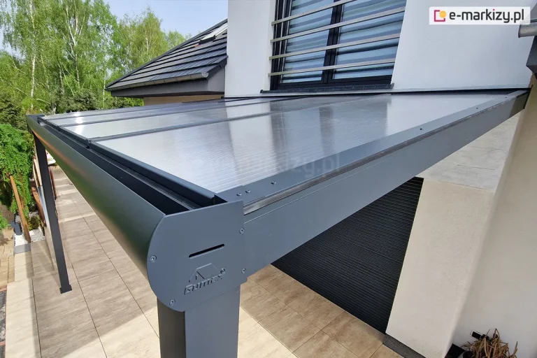 Narożnik zadaszenia tarasu w kolorze antracytowym z wypełnieniem z poliwęglanu komorowego. Zadaszenie z półokrągłą rynną zamocowane nad tarasem wyłożonym płytkami w kolorze brązowo-beżowym.