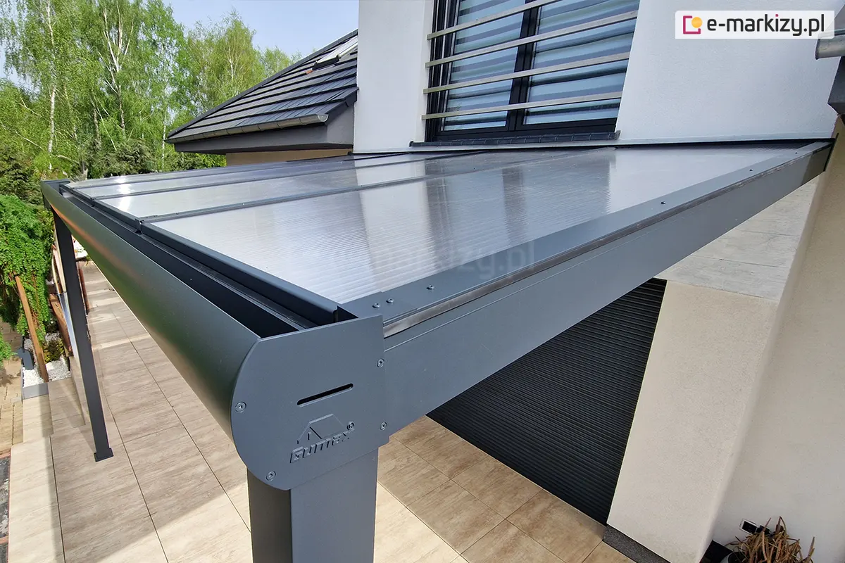 Narożnik zadaszenia tarasu w kolorze antracytowym z wypełnieniem z poliwęglanu komorowego. Zadaszenie z półokrągłą rynną zamocowane nad tarasem wyłożonym płytkami w kolorze brązowo-beżowym.