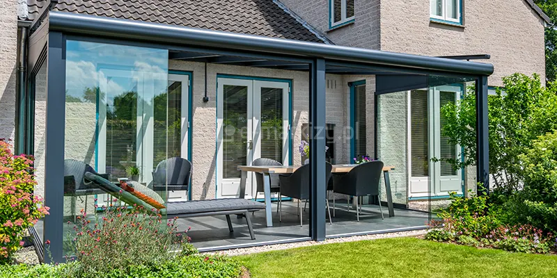 Przeszklone zadaszenie tarasu Terano, które zostało przekształcone w zamknięty ogród letni dzięki zastosowaniu przesuwnych systemów szklanych. Aluminiowa konstrukcja w kolorze antracytowym posiada charakterystyczną, zaokrąglową rynnę czołową, która płynnie łączy się z pionowymi słupami nośnymi. Ściany boczne oraz frontowe wypełniają bezramowe panele szklane, które można przesuwać po wielotorowych prowadnicach, co pozwala na całkowite lub częściowe otwarcie przestrzeni na ogród. Zadaszenie jest zamontowane do jasnej, tynkowanej elewacji budynku jednorodzinnego z dachem krytym ciemną dachówką. Wewnątrz tak powstałego ogrodu zimowego znajduje się strefa jadalniana ze stołem o jasnym blacie i czarnymi fotelami oraz leżak wypoczynkowy z szarym materacem. Podłoże tarasu wyłożone jest ciemnymi, kwadratowymi płytami, a wokół konstrukcji widoczna jest opaska z jasnego żwiru. Całość otacza niska roślinność ogrodowa, krzewy oraz zadbany trawnik.