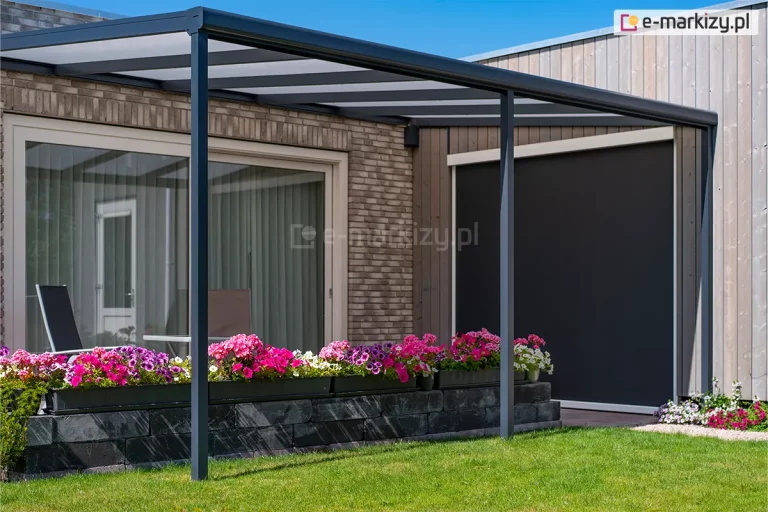 Zdjęcie prezentuje boczną perspektywę zadaszenia tarasowego Nebbiolo marki Deponti, zamontowanego przy elewacji łączącej jasną płytkę klinkierową z pionowymi panelami drewnianymi. Antracytowa konstrukcja z aluminium składa się z dachu opartego na krokwiach, rynny frontowej oraz pionowych słupów nośnych o kwadratowym przekroju. Wypełnienie dachu stanowią płyty poliwęglanu komorowego w kolorze mlecznym (opal), które zapewniają osłonę przed słońcem przy zachowaniu jasności na tarasie. Pod zadaszeniem widoczne są duże przeszklenia drzwi przesuwnych oraz zamontowana na sąsiedniej ścianie pionowa roleta typu screen w kolorze czarnym, chroniąca przed słońcem i wiatrem. Granicę tarasu wyznacza murek z ciemnych bloczków betonowych, na którym ustawiono długie donice z obficie kwitnącymi kwiatami w odcieniach intensywnego różu i fioletu. W tle widać fragment zielonego trawnika oraz bezchmurne, błękitne niebo.