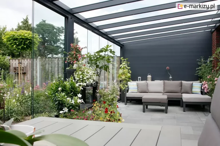 Widok z wnętrza nowoczesnego ogrodu letniego opartego na systemie zadaszenia tarasowego oraz bocznych, bezramowych ścian szklanych Auris. Zdjęcie prezentuje przestronne wnętrze z dachem jednospadowym o konstrukcji aluminiowej w kolorze antracytowym, z wypełnieniem z przezroczystego szkła bezpiecznego. Ściany boczne stanowią panoramiczne, bezramowe tafle szkła hartowanego, które zapewniają niezakłócony widok na bogato urządzony ogród z licznymi roślinami ozdobnymi, kwiatami oraz drewnianym ogrodzeniem. Wewnątrz znajduje się strefa relaksu z dużą, narożną sofą w kolorze szarym, ozdobioną pasiastymi poduszkami, oraz pasujący podnóżek na jasnoszarych płytach tarasowych. Tylna ściana konstrukcji jest pełna, wykończona gładkim panelem w kolorze ciemnego grafitu.