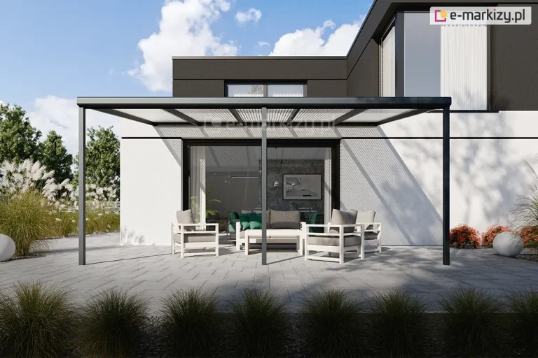 Zdjęcie prezentuje wizualizację nowoczesnej strefy wypoczynkowej na przestronnym tarasie, osłoniętym zadaszeniem Nebbiolo marki Deponti. Konstrukcja z matowego aluminium w kolorze antracytowym składa się z dachu wspartego na trzech pionowych słupach o kwadratowym przekroju, z których jeden znajduje się w centralnej części frontu. Dach posiada wypełnienie z przezroczystego poliwęglanu komorowego, który rzuca wyraźny, liniowy cień na białą elewację budynku. Pod zadaszeniem, na jasnoszarej nawierzchni z dużych płyt betonowych, ustawiono zestaw luksusowych mebli ogrodowych w kolorze białym z szarymi poduszkami, obejmujący sofę, dwa fotele oraz stolik kawowy. W tle widoczny jest minimalistyczny, piętrowy dom o biało-czarnej elewacji z dużymi przeszkleniami. Otoczenie tarasu wypełniają ozdobne trawy, niskie krzewy z czerwonymi kwiatami oraz białe, kuliste dekoracje ogrodowe pod jasnym niebem z rozproszonymi chmurami.