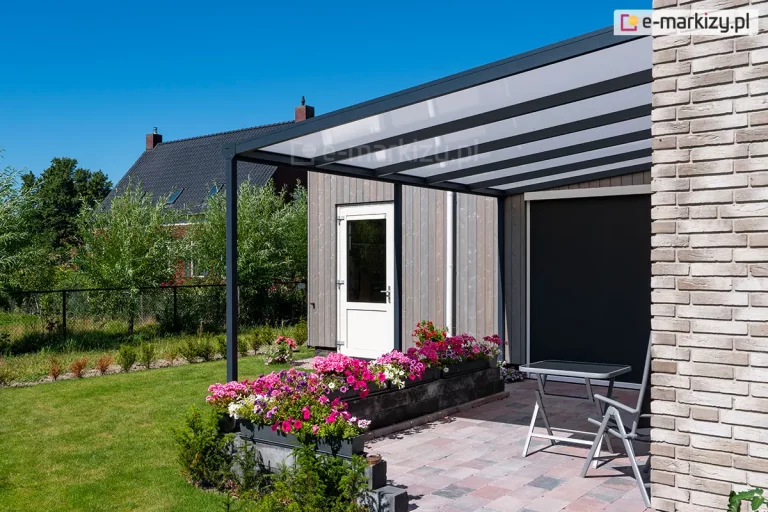 Zdjęcie prezentuje boczne ujęcie zadaszenia tarasowego Nebbiolo marki Deponti, zamontowanego do elewacji budynku wyłożonej jasną, beżowo-brązową cegłą klinkierową. Konstrukcja zadaszenia wykonana jest z profili aluminiowych w kolorze antracytowym, a jej dach wypełniają płyty poliwęglanu komorowego o mlecznym wybarwieniu, ułożone rzędowo między krokwiami. Pod zadaszeniem, na wybrukowanym kostką tarasie, widoczne są szare meble ogrodowe, w tym krzesło i stolik z szklanym blatem, a obok nich pionowa roleta zewnętrzna typu screen w ciemnym kolorze. Krawędź tarasu wyznacza murek z czarnych, łupanych bloczków betonowych, na którym ustawiono rządek donic z gęsto kwitnącymi pelargoniami i petuniami w kolorach fioletowym, różowym oraz białym. W tle rozpościera się widok na zielony ogród z drzewami oraz sąsiednie domy jednorodzinne o spadzistych, ciemnych dachach, widoczne pod bezchmurnym, intensywnie błękitnym niebem.