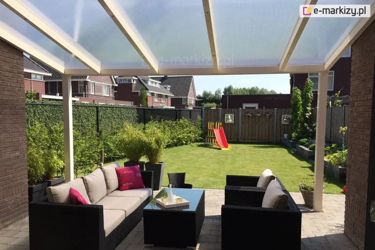 Widok spod nowoczesnego zadaszenia tarasowego Lunaro o jasnej, kremowej konstrukcji, która została zamontowana przy budynku z brązowej cegły. Jednospadowy dach posiada wypełnienie z przezroczystych paneli poliwęglanu komorowego, wspartych na smukłych krokwiach i solidnych słupach o kwadratowym przekroju. Pod zadaszeniem, na tarasie wyłożonym szarą kostką brukową, zaaranżowano strefę wypoczynkową z dużym zestawem mebli z czarnego technorattanu, obejmującym długą sofę z jasnoszarymi i różowymi poduszkami, dwa fotele oraz stolik kawowy z ciemnym, szklanym blatem. Wzdłuż lewej oraz części frontowej krawędzi tarasu ustawiono wysokie, grafitowe donice z gęstymi, zielonymi krzewami. W tle rozpościera się widok na zadbany, słoneczny ogród z zielonym trawnikiem, na którym znajduje się kolorowa zjeżdżalnia dla dzieci. Posesja jest ogrodzona siatką oraz drewnianym płotem, za którymi widoczne są dachy sąsiednich domów jednorodzinnych pod czystym, błękitnym niebem.