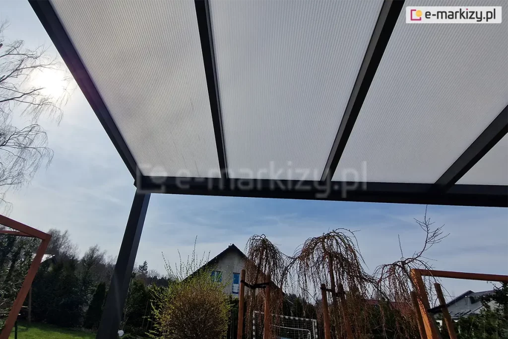 Zadaszenie z poliwęglanem komorowym Modes ustawione na tarasie z widokiem na dom sąsiada oraz drzewka ustawione przy tarasie. W tle widoczne także szaroniebieskie niebo wczesnej wiosny, z którego przenika kilka promieni słońca.