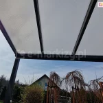 Zadaszenie z poliwęglanem komorowym Modes ustawione na tarasie z widokiem na dom sąsiada oraz drzewka ustawione przy tarasie. W tle widoczne także szaroniebieskie niebo wczesnej wiosny, z którego przenika kilka promieni słońca.