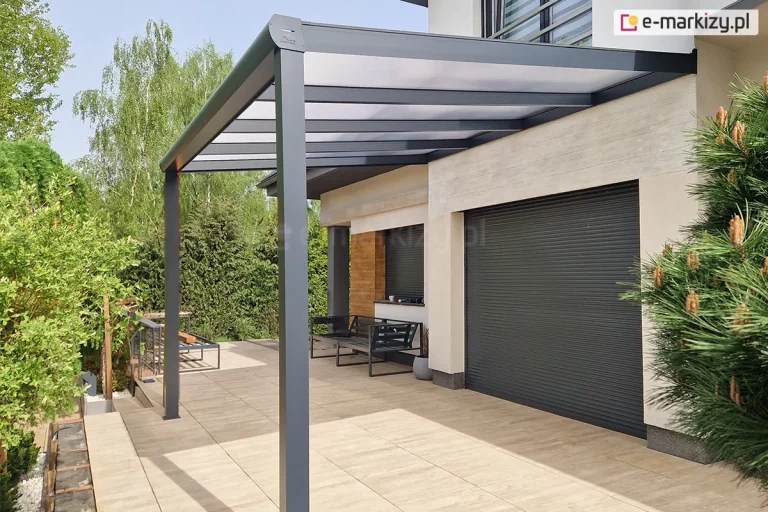 Nowoczesne zadaszenie tarasowe wykonane z antracytowych profili aluminiowych, które jest przytwierdzone bezpośrednio do jasnej elewacji piętrowego budynku mieszkalnego. Konstrukcja dachu posiada wypełnienie z mlecznych, półprzezroczystych paneli poliwęglanowych, które są równomiernie rozmieszczone pomiędzy poprzecznymi krokwiami. Całość opiera się na dwóch solidnych, kwadratowych słupach nośnych usytuowanych na krawędzi tarasu wyłożonego dużymi, beżowymi płytami gresowymi. Pod zadaszeniem, przy ścianie z dużą, szarą roletą zewnętrzną, ustawiono prostą czarną ławkę o metalowej konstrukcji, a obok widoczna jest ozdobna wstawka elewacyjna o strukturze drewna. Otoczenie tarasu stanowi gęsta ściana zielonych krzewów oraz wysokie brzozy w tle, a na pierwszym planie po prawej stronie widać fragment gałęzi sosny z młodymi przyrostami.