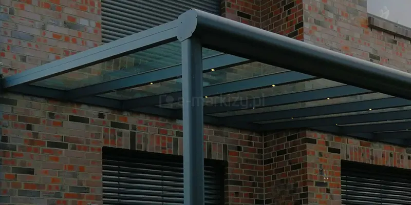 Techniczne zbliżenie na detal konstrukcyjny aluminiowego zadaszenia tarasu Terano w kolorze antracytowym. Widoczny jest punkt styku pionowego słupa nośnego o kwadratowym przekroju z przednią belką rynnową o charakterystycznym, zaokrąglonym kształcie. Belka rynnowa jest wykończona dopasowaną estetycznie zaślepką boczną montowaną na śruby. Konstrukcja dachu opiera się na równolegle rozmieszczonych krokwiach aluminiowych, w których zamontowano zintegrowane, punktowe oświetlenie LED w formie małych, okrągłych oczek. Wypełnienie dachu stanowią przezroczyste panele szklane, które są widoczne nad profilami krokwi. Tło stanowi elewacja budynku z czerwono-brązowej cegły klinkierowej oraz ciemne żaluzje fasadowe w oknach. Fotografia akcentuje precyzję łączeń elementów konstrukcyjnych oraz funkcjonalność systemowego oświetlenia tarasowego.