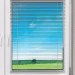 Białe okno z PCV wyposażone w srebrne żaluzje poziome aluminiowe 16 mm, zamontowane bezinwazyjnie na ramie okiennej. Żaluzje mają cienkie, lekko lśniące lamele, które są ustawione w pozycji otwartej, umożliwiając widok na zewnątrz. Przez szybę widoczny jest sielski krajobraz z intensywnie zielonym polem, pojedynczym drzewem pośrodku oraz bezchmurnym, jasnoniebieskim niebem. Konstrukcja żaluzji obejmuje górną rynnę montażową oraz dolną belkę obciążającą, połączone systemem cienkich sznurków drabinkowych. Po prawej stronie u góry znajduje się przezroczyste pokrętło służące do regulacji kąta nachylenia lameli oraz sznurek do podciągania osłony. Rama okna jest biała, o gładkiej powierzchni, z widoczną klamką po lewej stronie oraz zawiasem po prawej. Ściana wokół okna wykończona jest tapetą o delikatnym, geometrycznym wzorze składającym się z drobnych, jasnych linii tworzących romby lub koła na szarym tle. Całość kompozycji jest jasna i prezentuje produkt w naturalnych warunkach oświetleniowych.