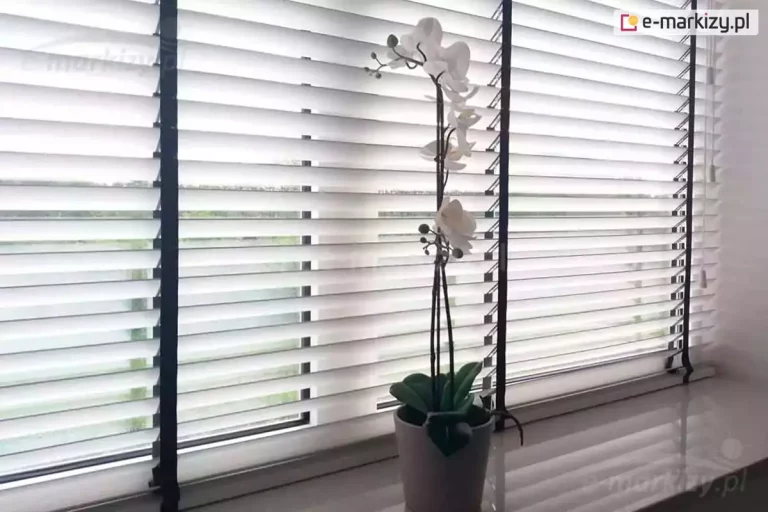 Zdjęcie prezentuje biała żaluzja aluminiowa 25mm zamontowana na dużym oknie, tworzące nowoczesną i jasną aranżację wnętrza. Lamele o szerokości dwudziestu pięciu milimetrów są ustawione w pozycji poziomej, lekko uchylonej, co pozwala na subtelne przenikanie światła dziennego do pomieszczenia. Charakterystycznym elementem tej osłony okiennej są szerokie, czarne taśmy drabinkowe, które stanowią mocny akcent dekoracyjny i kontrastują z czystą bielą aluminium. Na jasnym parapecie, na tle żaluzji, stoi doniczka z białym storczykiem (orchideą), którego pionowa łodyga z kwiatami i pąkami harmonizuje z pionowymi pasami drabinek. Przez szczeliny między lamelami widać zarys zewnętrznego krajobrazu z zielenią drzew. Po prawej stronie widoczne są białe sznurki służące do podnoszenia i opuszczania żaluzji, zakończone estetycznymi obciążnikami. Całość kompozycji jest utrzymana w minimalistycznym stylu, podkreślając funkcjonalność i elegancję żaluzji aluminiowych.