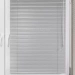 Zawieszona bezinwazyjnie na białej ramie skrzydła okna żaluzja aluminiowa 25 mm z lamelami w jasnoszarym, perłowym odcieniu. W pełni zamknięte lamele przesłaniają całą szybę. Po prawej stronie żaluzji widoczne elementy sterujące: sznurek do podciągania i opuszczania lamel oraz pokrętło lexanowe do obracania lamelek.