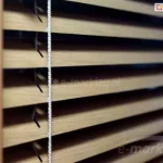 Żaluzja aluminiowa 25 mm drewnopodobna okleina lekko uchylonych lameli z biała drabinka sznurkowa tworzą ciepłą osłonę na okno.