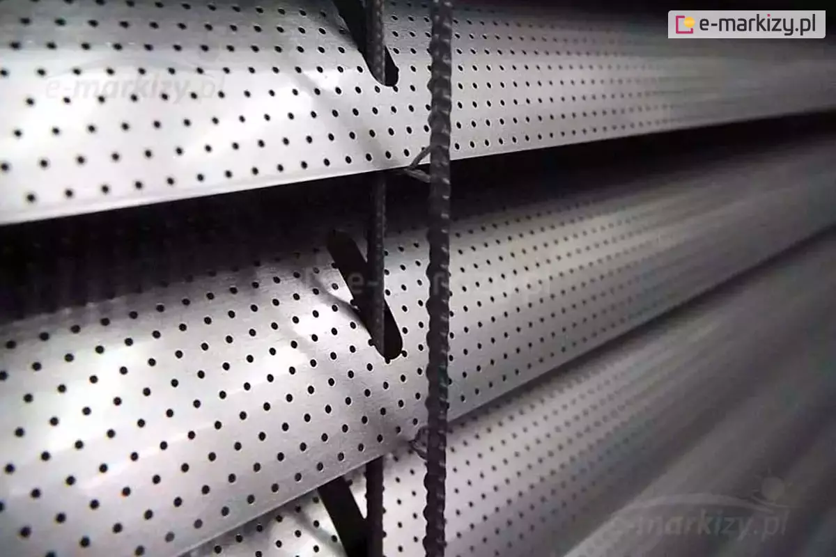 Szczegółowe zbliżenie na lamele poziomej żaluzji fasadowej wykonanej z aluminium w kolorze srebrnym lub jasnoszarym. Powierzchnia lameli posiada regularną perforację w postaci drobnych, gęsto rozmieszczonych otworów, co pozwala na częściowe przenikanie światła i powietrza przy jednoczesnym zachowaniu ochrony przed słońcem. Lamele mają lekko wygięty, wypukły profil zwiększający ich sztywność konstrukcyjną. Przez owalne wycięcia wzdłuż krawędzi lameli przechodzą pionowe elementy sterujące w postaci tekstylnych linek w kolorze ciemnoszarym, które są połączone systemem drabinkowym utrzymującym lamele w równych odstępach. Mechanizm ten umożliwia regulację kąta nachylenia lameli, co pozwala na precyzyjne sterowanie ilością światła wpadającego do pomieszczenia. Oświetlenie na zdjęciu podkreśla metaliczną teksturę materiału oraz techniczną precyzję wykonania perforacji.
