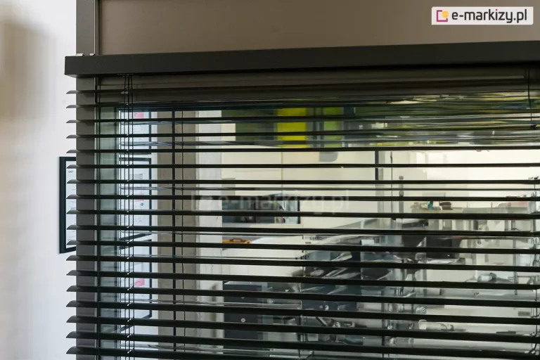 Zbliżenie na górną sekcję żaluzji poziomej C50 Slim zamontowanej wewnątrz przeszklonego pomieszczenia biurowego lub technicznego. Zdjęcie prezentuje czarne lamele o delikatnie wygiętym profilu, które są zawieszone na systemie drabinek sznurkowych, zapewniających ich równomierne rozmieszczenie i możliwość regulacji kąta nachylenia. Górna część mechanizmu jest osłonięta solidną, ciemnobrązową maskownicą o matowym wykończeniu, która integruje się z konstrukcją ramy okiennej. Przez przezroczyste przeszklenie widoczne jest tło z elementami wyposażenia warsztatowego lub magazynowego, a po lewej stronie na białej ścianie zawieszona jest ramka z dokumentem lub certyfikatem. Detal ukazuje precyzję wykonania elementów sterujących oraz sposób montażu systemu żaluzji przy samej krawędzi górnego profilu.