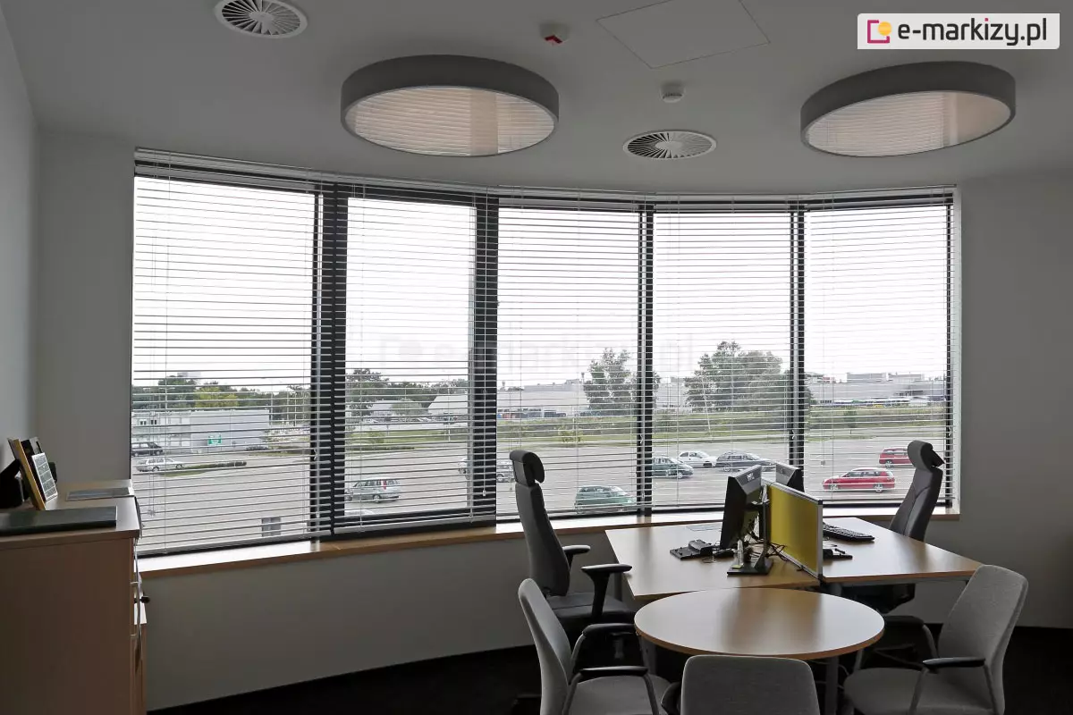 Widok z wnętrza nowoczesnego biura na panoramiczne, łukowe przeszklenie wyposażone w system wewnętrznych żaluzji poziomych C50 Slim. Srebrne, aluminiowe lamele są ustawione w pozycji otwartej, co zapewnia optymalną kontrolę nad dopływem światła dziennego przy zachowaniu widoczności na zewnętrzny parking oraz infrastrukturę miejską. W pomieszczeniu znajduje się stanowisko pracy z biurkiem o jasnym blacie, wyposażone w monitory komputerowe oddzielone żółtym panelem akustycznym oraz ergonomiczne fotele biurowe. Na suficie zamontowano dwie duże, okrągłe oprawy oświetleniowe typu plafon oraz okrągłe anemostaty systemu wentylacji. Estetyka wnętrza jest minimalistyczna, z białymi ścianami i ciemną wykładziną, co podkreśla techniczny charakter zastosowanych osłon okiennych dopasowanych do krzywizny fasady.