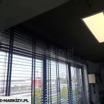 Zdjęcie przedstawia nowoczesne wnętrze biurowe lub usługowe w porze wieczornej lub przy przygaszonym oświetleniu, gdzie głównym elementem są żaluzje drewniane 25 mm w kolorze ciemnego antracytu lub czerni. Przesłona pokrywa szerokie, panoramiczne przeszklenie podzielone pionowymi profilami. Żaluzje są opuszczone, a ich lamele o szerokości 25 mm są ustawione w pozycji poziomej, co pozwala na wgląd w oświetloną infrastrukturę zewnętrzną – widoczne są budynki handlowe, parking oraz zieleń miejska pod ciemniejącym niebem. Konstrukcja żaluzji opiera się na systemie drabinek sznurkowych, które przebiegają pionowo przez całą płaszczyznę osłony. Sufit pomieszczenia jest ciemny, prawdopodobnie wykończony w surowym, industrialnym stylu, a zamontowana na nim prostokątna lampa panelowa emituje intensywne, ciepłe światło, które kontrastuje z chłodnym odcieniem wpadającym przez okno. Po prawej stronie kadru widoczny jest fragment ściany z fototapetą przedstawiającą motyw lasu w skali szarości oraz czarna, wisząca lampa o kopułowatym kształcie. Żaluzje drewniane 25 mm idealnie komponują się z nowoczesnym, minimalistycznym designem wnętrza, podkreślając jego techniczny i elegancki charakter.