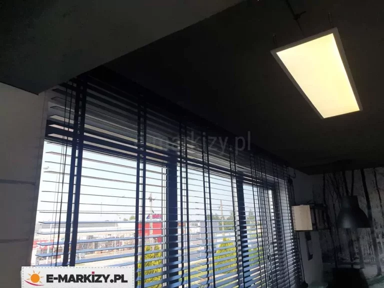 Zdjęcie przedstawia nowoczesne wnętrze biurowe lub usługowe w porze wieczornej lub przy przygaszonym oświetleniu, gdzie głównym elementem są żaluzje drewniane 25 mm w kolorze ciemnego antracytu lub czerni. Przesłona pokrywa szerokie, panoramiczne przeszklenie podzielone pionowymi profilami. Żaluzje są opuszczone, a ich lamele o szerokości 25 mm są ustawione w pozycji poziomej, co pozwala na wgląd w oświetloną infrastrukturę zewnętrzną – widoczne są budynki handlowe, parking oraz zieleń miejska pod ciemniejącym niebem. Konstrukcja żaluzji opiera się na systemie drabinek sznurkowych, które przebiegają pionowo przez całą płaszczyznę osłony. Sufit pomieszczenia jest ciemny, prawdopodobnie wykończony w surowym, industrialnym stylu, a zamontowana na nim prostokątna lampa panelowa emituje intensywne, ciepłe światło, które kontrastuje z chłodnym odcieniem wpadającym przez okno. Po prawej stronie kadru widoczny jest fragment ściany z fototapetą przedstawiającą motyw lasu w skali szarości oraz czarna, wisząca lampa o kopułowatym kształcie. Żaluzje drewniane 25 mm idealnie komponują się z nowoczesnym, minimalistycznym designem wnętrza, podkreślając jego techniczny i elegancki charakter.