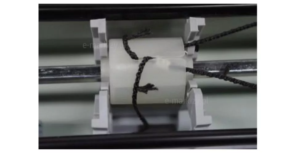Zdjęcie przedstawia zbliżenie na wewnętrzny mechanizm sterujący, który obsługuje żaluzje drewniane 50 mm, umieszczony wewnątrz stalowej rynny górnej o czarnym wykończeniu. Centralnym punktem fotografii jest biała, plastikowa rolka prowadząca osadzona na metalowym, srebrnym pręcie obrotowym o przekroju kwadratowym lub sześciokątnym, który przechodzi poziomo przez całą szerokość mechanizmu. Na rolkę nawinięte są czarne, plecione sznurki drabinki, które odpowiadają za podnoszenie, opuszczanie oraz regulację kąta nachylenia lameli w systemie żaluzje drewniane 50 mm. Rolka jest zabezpieczona z obu stron przez szare, polimerowe wsporniki stabilizujące, które utrzymują ją w odpowiedniej pozycji i zapobiegają przesuwaniu się wzdłuż osi pręta. Widoczne są końcówki sznurków z charakterystycznym splotem, przechodzące przez szczeliny w mechanizmie, co pozwala na precyzyjne sterowanie osłoną okienną. Całość konstrukcji ukazuje techniczne aspekty budowy napędu, podkreślając solidność komponentów użytych do produkcji żaluzji o szerokich lamelach.