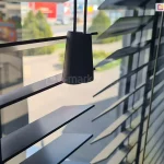 Zdjęcie prezentuje zbliżenie na żaluzje drewniane 50 mm w eleganckim, czarnym kolorze o satynowym wykończeniu, zamontowane w świetle okna. Na pierwszym planie widoczne są dwa czarne, drewniane dzwonki o stożkowym kształcie, które stanowią estetyczne zakończenie sznurków operacyjnych służących do podnoszenia i opuszczania osłony. Lamele, z których składają się żaluzje drewniane 50 mm, są ustawione pod skosem w pozycji półotwartej, co pozwala na subtelne przenikanie światła dziennego do wnętrza i jednoczesne zachowanie prywatności. Przez szczeliny między lamelami prześwituje rozmyte tło przedstawiające widok z okna na infrastrukturę miejską oraz zieleń. Konstrukcja wykorzystuje klasyczne prowadzenie sznurkowe, gdzie cienkie, czarne nitki drabinki przechodzą pionowo przez krawędzie lameli, zapewniając ich równomierne rozłożenie. Fotografia podkreśla minimalistyczny design produktu oraz wysoką jakość detali wykończeniowych, które idealnie komponują się z nowoczesnymi aranżacjami wnętrz.