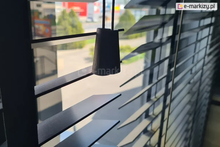 Zdjęcie prezentuje zbliżenie na żaluzje drewniane 50 mm w eleganckim, czarnym kolorze o satynowym wykończeniu, zamontowane w świetle okna. Na pierwszym planie widoczne są dwa czarne, drewniane dzwonki o stożkowym kształcie, które stanowią estetyczne zakończenie sznurków operacyjnych służących do podnoszenia i opuszczania osłony. Lamele, z których składają się żaluzje drewniane 50 mm, są ustawione pod skosem w pozycji półotwartej, co pozwala na subtelne przenikanie światła dziennego do wnętrza i jednoczesne zachowanie prywatności. Przez szczeliny między lamelami prześwituje rozmyte tło przedstawiające widok z okna na infrastrukturę miejską oraz zieleń. Konstrukcja wykorzystuje klasyczne prowadzenie sznurkowe, gdzie cienkie, czarne nitki drabinki przechodzą pionowo przez krawędzie lameli, zapewniając ich równomierne rozłożenie. Fotografia podkreśla minimalistyczny design produktu oraz wysoką jakość detali wykończeniowych, które idealnie komponują się z nowoczesnymi aranżacjami wnętrz.