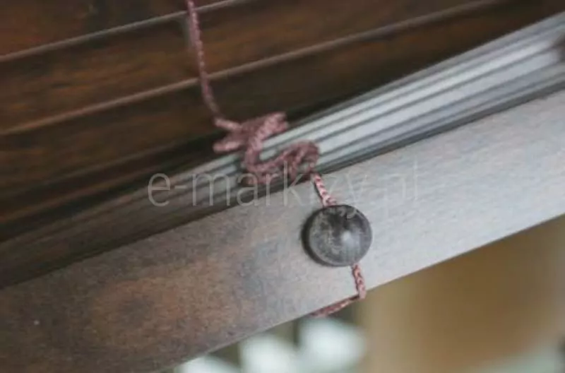 Zdjęcie prezentuje zbliżenie na dolny wykończeniowy element konstrukcji, jaką są żaluzje drewniane 50 mm w kolorze ciemnego, egzotycznego drewna o wyraźnym usłojeniu. Na pierwszym planie widoczna jest masywna belka dolna żaluzji, w której osadzony został okrągły, wypukły guziczek maskujący wykonany z tworzywa lub drewna w dopasowanym, ciemnobrązowym odcieniu. Przez otwór w belce przechodzi cienki, pleciony sznurek drabinki sznurkowej w kolorze brązowym, który stabilizuje żaluzje drewniane 50 mm i odpowiada za ich pionowe prowadzenie. Sznurek tworzy charakterystyczną pętlę i przechodzi przez kolejne lamele widoczne w górnej części kadru, które są ułożone równolegle względem siebie. Perspektywa zdjęcia pod kątem pozwala dostrzec matowe wykończenie powierzchni drewna oraz precyzję montażu systemu sterowania sznurkowego. Tło jest lekko rozmyte, co skupia uwagę na detalu wykończenia dolnej krawędzi osłony okiennej oraz sposobie zabezpieczenia zakończenia drabinki.
