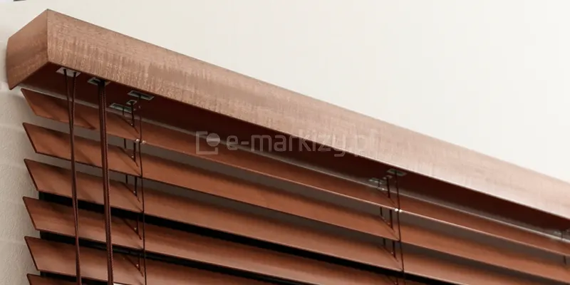 Zdjęcie prezentuje górną część systemu, jakim są żaluzje drewniane 50 mm, skupiając się na estetycznym wykończeniu maskownicy rynny górnej. Maskownica, wykonana z litego drewna w ciepłym odcieniu średniego brązu (np. dąb lub orzech), posiada profilowany kształt z wyraźnie zaznaczoną górną krawędzią, co nadaje osłonie klasyczny wygląd. Pod drewnianym gzymsem widoczne są precyzyjnie ułożone lamele o szerokości 50 mm, które są połączone za pomocą cienkich, brązowych sznurków drabinki sznurkowej. Sznurki te przechodzą przez metalowe, prostokątne przelotki zamontowane w dolnej części rynny, co zapewnia płynną pracę mechanizmu podnoszenia i obracania listew. Po lewej stronie kadru zwisają cztery sznurki operacyjne w kolorze dopasowanym do drewna, służące do sterowania poziomem zaciemnienia wnętrza. Całość konstrukcji żaluzje drewniane 50 mm jest zamontowana blisko ściany o jasnej, chropowatej fakturze, co podkreśla naturalną kolorystykę i strukturę użytego drewna.