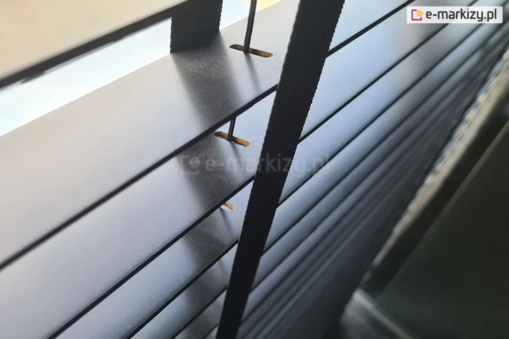 Zdjęcie prezentuje zbliżenie na żaluzje drewniane 50 mm w głębokim, czarnym kolorze o eleganckim, matowym wykończeniu. Perspektywa z góry pozwala na dokładne przyjrzenie się strukturze i komponentom systemu sterowania. Głównym elementem kadru jest pionowa, czarna drabinka taśmowa o gęstym splocie, która przebiega prostopadle do poziomych lameli. Tuż obok taśmy widoczny jest cienki, czarny sznurek prowadzący, który przechodzi przez precyzyjnie wyfrezowane, podłużne otwory w lamelach. Wnętrze otworów ukazuje naturalną, jasną strukturę drewna, co stanowi subtelny kontrast dla czarnej powłoki zewnętrznej, jaką pokryte są żaluzje drewniane 50 mm. Lamele są ustawione równolegle w pozycji otwartej, odsłaniając jasne, rozproszone światło wpadające z zewnątrz, które delikatnie odbija się od krawędzi listew. Fotografia techniczna podkreśla minimalistyczną estetykę produktu oraz wysoką jakość dopasowania kolorystycznego wszystkich elementów osprzętu, w tym drabinek i sznurków, do ciemnej kolorystyki drewna.