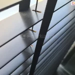 Zdjęcie prezentuje zbliżenie na żaluzje drewniane 50 mm w głębokim, czarnym kolorze o eleganckim, matowym wykończeniu. Perspektywa z góry pozwala na dokładne przyjrzenie się strukturze i komponentom systemu sterowania. Głównym elementem kadru jest pionowa, czarna drabinka taśmowa o gęstym splocie, która przebiega prostopadle do poziomych lameli. Tuż obok taśmy widoczny jest cienki, czarny sznurek prowadzący, który przechodzi przez precyzyjnie wyfrezowane, podłużne otwory w lamelach. Wnętrze otworów ukazuje naturalną, jasną strukturę drewna, co stanowi subtelny kontrast dla czarnej powłoki zewnętrznej, jaką pokryte są żaluzje drewniane 50 mm. Lamele są ustawione równolegle w pozycji otwartej, odsłaniając jasne, rozproszone światło wpadające z zewnątrz, które delikatnie odbija się od krawędzi listew. Fotografia techniczna podkreśla minimalistyczną estetykę produktu oraz wysoką jakość dopasowania kolorystycznego wszystkich elementów osprzętu, w tym drabinek i sznurków, do ciemnej kolorystyki drewna.