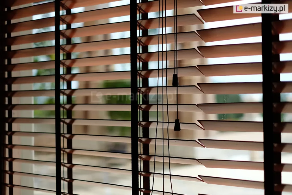 Zdjęcie prezentuje zbliżenie na żaluzje drewniane 50 mm w kolorze naturalnego, jasnego drewna, zamontowane w oknie i ustawione w pozycji poziomej (otwartej). Głównym elementem kompozycji są trzy ciemnobrązowe drabinki taśmowe o szerokim splocie, które przebiegają pionowo przez całą wysokość osłony, stabilizując lamele i nadając im nowoczesny kontrastowy wygląd. Przez środek prawej części kadru przebiegają cztery cienkie, czarne sznurki operacyjne, które kończą się na różnych wysokościach dwoma ozdobnymi dzwonkami o stożkowym kształcie, wykonanymi z czarnego tworzywa lub malowanego drewna. Żaluzje drewniane 50 mm są ukazane na tle jasnego, rozmytego widoku zewnętrznego, co podkreśla ich funkcję regulacji dopływu światła do pomieszczenia. Lamele mają gładką powierzchnię i precyzyjnie docięte krawędzie, a ich równomierne rozstawienie demonstruje wysoką jakość mechanizmu sterującego. Światło dzienne wpadające między listwami tworzy rytmiczne pasy, eksponując techniczne detale montażu drabinek taśmowych.