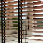 Zdjęcie prezentuje zbliżenie na żaluzje drewniane 50 mm w kolorze naturalnego, jasnego drewna, zamontowane w oknie i ustawione w pozycji poziomej (otwartej). Głównym elementem kompozycji są trzy ciemnobrązowe drabinki taśmowe o szerokim splocie, które przebiegają pionowo przez całą wysokość osłony, stabilizując lamele i nadając im nowoczesny kontrastowy wygląd. Przez środek prawej części kadru przebiegają cztery cienkie, czarne sznurki operacyjne, które kończą się na różnych wysokościach dwoma ozdobnymi dzwonkami o stożkowym kształcie, wykonanymi z czarnego tworzywa lub malowanego drewna. Żaluzje drewniane 50 mm są ukazane na tle jasnego, rozmytego widoku zewnętrznego, co podkreśla ich funkcję regulacji dopływu światła do pomieszczenia. Lamele mają gładką powierzchnię i precyzyjnie docięte krawędzie, a ich równomierne rozstawienie demonstruje wysoką jakość mechanizmu sterującego. Światło dzienne wpadające między listwami tworzy rytmiczne pasy, eksponując techniczne detale montażu drabinek taśmowych.