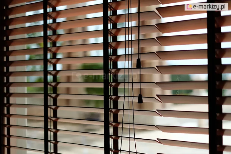 Zdjęcie prezentuje zbliżenie na żaluzje drewniane 50 mm w kolorze naturalnego, jasnego drewna, zamontowane w oknie i ustawione w pozycji poziomej (otwartej). Głównym elementem kompozycji są trzy ciemnobrązowe drabinki taśmowe o szerokim splocie, które przebiegają pionowo przez całą wysokość osłony, stabilizując lamele i nadając im nowoczesny kontrastowy wygląd. Przez środek prawej części kadru przebiegają cztery cienkie, czarne sznurki operacyjne, które kończą się na różnych wysokościach dwoma ozdobnymi dzwonkami o stożkowym kształcie, wykonanymi z czarnego tworzywa lub malowanego drewna. Żaluzje drewniane 50 mm są ukazane na tle jasnego, rozmytego widoku zewnętrznego, co podkreśla ich funkcję regulacji dopływu światła do pomieszczenia. Lamele mają gładką powierzchnię i precyzyjnie docięte krawędzie, a ich równomierne rozstawienie demonstruje wysoką jakość mechanizmu sterującego. Światło dzienne wpadające między listwami tworzy rytmiczne pasy, eksponując techniczne detale montażu drabinek taśmowych.