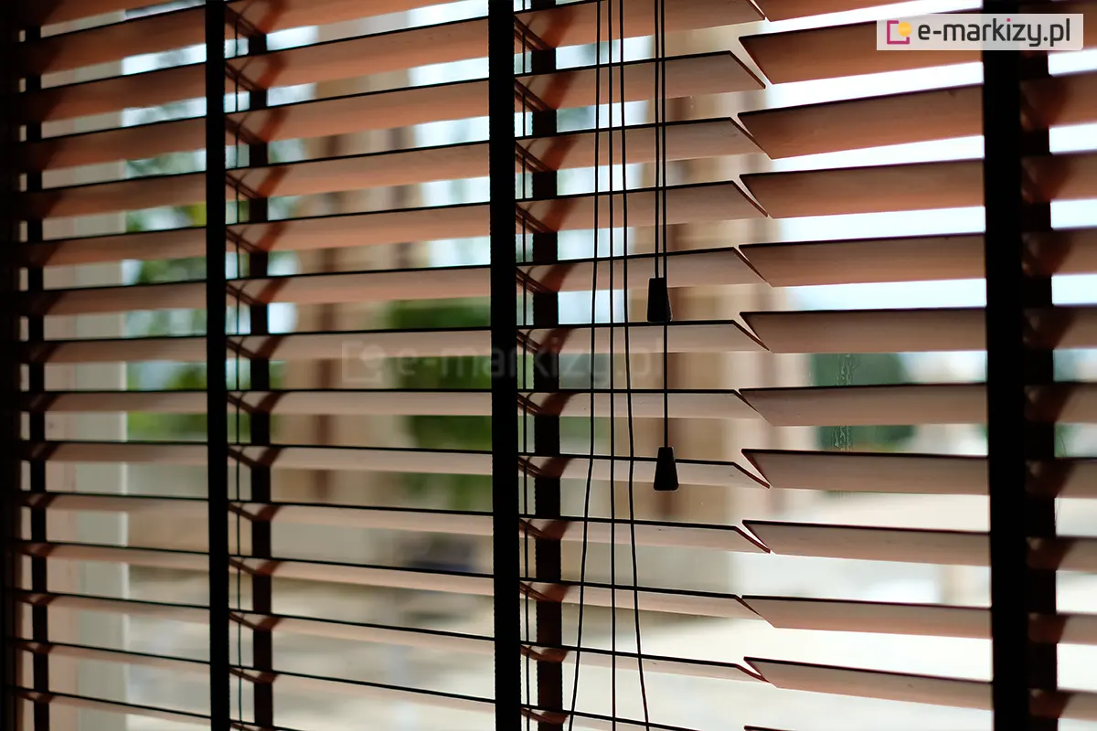 Zdjęcie prezentuje zbliżenie na żaluzje drewniane 50 mm w kolorze naturalnego, jasnego drewna, zamontowane w oknie i ustawione w pozycji poziomej (otwartej). Głównym elementem kompozycji są trzy ciemnobrązowe drabinki taśmowe o szerokim splocie, które przebiegają pionowo przez całą wysokość osłony, stabilizując lamele i nadając im nowoczesny kontrastowy wygląd. Przez środek prawej części kadru przebiegają cztery cienkie, czarne sznurki operacyjne, które kończą się na różnych wysokościach dwoma ozdobnymi dzwonkami o stożkowym kształcie, wykonanymi z czarnego tworzywa lub malowanego drewna. Żaluzje drewniane 50 mm są ukazane na tle jasnego, rozmytego widoku zewnętrznego, co podkreśla ich funkcję regulacji dopływu światła do pomieszczenia. Lamele mają gładką powierzchnię i precyzyjnie docięte krawędzie, a ich równomierne rozstawienie demonstruje wysoką jakość mechanizmu sterującego. Światło dzienne wpadające między listwami tworzy rytmiczne pasy, eksponując techniczne detale montażu drabinek taśmowych.