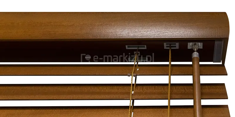 Zdjęcie prezentuje techniczne zbliżenie na górną sekcję produktu, jakim są żaluzje drewniane 50 mm w kolorze nasyconego brązu o wyraźnej strukturze drewna. Centralnym elementem jest drewniana maskownica o profilowanym, zaokrąglonym kształcie, która osłania stalową rynnę górną. Od spodu rynny widoczne są trzy różne porty techniczne wykonane z szarego tworzywa: prostokątny otwór, przez który przechodzą sznurki drabinki, kwadratowy przepust dla sznurków operacyjnych oraz metalowy zaczep z przegubem, na którym zamontowany jest drewniany drążek do regulacji kąta nachylenia listew. Lamele, z których zbudowane są żaluzje drewniane 50 mm, są ustawione poziomo, ukazując precyzyjne dopasowanie kolorystyczne sznurków drabinki do odcienia drewna. Drążek sterujący ma cylindryczny kształt i jest zakończony metalową skuwką z oczkiem, co umożliwia jego swobodny obrót i zmianę położenia lameli. Całość konstrukcji jest przedstawiona na białym, neutralnym tle, co pozwala wyeksponować detale mechaniczne oraz jakość wykończenia powierzchni drewnianych.