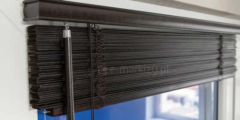 Zdjęcie prezentuje zbliżenie na górną część systemu osłonowego, jakim są żaluzje drewniane 25 mm w kolorze ciemnego wenge lub głębokiego brązu. Żaluzje są całkowicie podciągnięte do góry, tworząc zwarty pakiet poziomo ułożonych lameli tuż pod rynną górną. Rynna jest osłonięta ozdobną maskownicą wykonaną z drewna o tej samej barwie i subtelnym połysku, która posiada profilowane, lekko zaokrąglone krawędzie. Po lewej stronie widoczne jest pokrętło w formie sztywnego drążka o ziarnistej teksturze, służące do regulacji kąta nachylenia listewek. System wykorzystuje klasyczne drabinki sznurkowe, które przy podciągniętej żaluzji układają się w dekoracyjne, zygzakowate sploty widoczne na tle pakietu lameli. Dodatkowo widoczne są cienkie sznurki operacyjne służące do podnoszenia i opuszczania konstrukcji. Żaluzje drewniane 25 mm są zamontowane wewnątrz wnęki okiennej na białej ramie, co tworzy wyraźny kontrast kolorystyczny. W dolnej części kadru, pod pakietem drewna, prześwituje fragment szyby, przez którą wpada dzienne światło, odbijające się od krawędzi poszczególnych listewek i podkreślające ich gęste ułożenie.