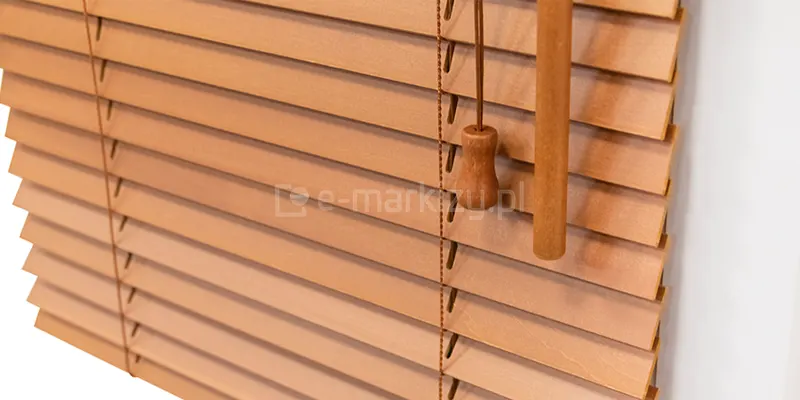 Zdjęcie prezentuje zbliżenie na żaluzje drewniane 50 mm w ciepłym, jasnobrązowym odcieniu, przypominającym naturalną olchę lub jasny dąb. Lamele są ustawione w pozycji lekko nachylonej, ukazując gładką strukturę materiału oraz precyzyjnie wycięte otwory na drabinkę sznurkową. Przez system przechodzi cienki, pleciony sznurek w kolorze brązowym, który stabilizuje żaluzje drewniane 50 mm i umożliwia ich podnoszenie. W prawej części kadru widoczne są elementy sterujące: drewniane zakończenie sznurka o ozdobnym kształcie dzwonka oraz fragment grubszego, drewnianego drążka służącego do regulacji kąta nachylenia listew. Produkty te charakteryzują się klasycznym designem, gdzie kolorystyka osprzętu, w tym sznurków i detali wykończeniowych, jest idealnie dopasowana do barwy drewna. Całość kompozycji na białym tle podkreśla estetykę i naturalny charakter osłony okiennej.