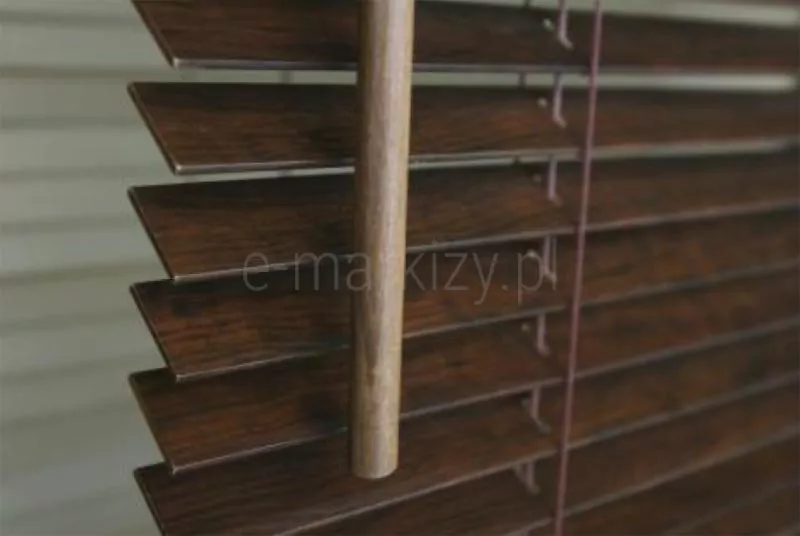 Zdjęcie prezentuje szczegółowe zbliżenie na żaluzje drewniane 25 mm w głębokim, ciemnobrązowym odcieniu z wyraźnie zarysowaną strukturą naturalnego usłojenia drewna. Lamele o szerokości 25 mm są ustawione poziomo w równych odstępach, a ich krawędzie są precyzyjnie wykończone i lekko matowe. Przez środek kadru przebiega pionowo drewniany drążek sterujący o cylindrycznym kształcie, służący do regulacji kąta nachylenia listewek. Drążek ma ten sam kolor i fakturę co lamele, co zapewnia spójność estetyczną całego systemu. W tle widoczna jest drobna drabinka sznurkowa, która stabilizuje poszczególne elementy żaluzji. Całość kompozycji jest ukazana w bliskim planie, co pozwala dostrzec detale faktury drewna oraz precyzję mechanizmu operacyjnego. Światło padające na żaluzje drewniane 25 mm podkreśla bogactwo ciemnej barwy i tworzy delikatne przejścia tonalne na powierzchni drewna.