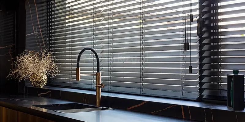 Zdjęcie prezentuje luksusową i nowoczesną aranżację kuchni, w której strefa zmywania została umieszczona bezpośrednio pod szerokim oknem przesłoniętym przez żaluzje drewniane 25 mm. Osłony okienne są w kolorze grafitowym lub bardzo ciemnym antracycie i posiadają matowe wykończenie. Lamele są ustawione w pozycji poziomej z lekkim nachyleniem, co zapewnia prywatność przy jednoczesnym wpuszczaniu pasm naturalnego światła do wnętrza. System sterowania żaluzjami znajduje się po prawej stronie i składa się z kilku cienkich sznurków zakończonych eleganckimi, podłużnymi obciążnikami w kolorze dopasowanym do drewna. Blat kuchenny oraz zintegrowany z nim zlewozmywak zostały wykonane z ciemnego konglomeratu lub kamienia o wyraźnym, jasnym żyłkowaniu przypominającym marmur. Na blacie zamontowano designerską baterię kuchenną w kolorze szczotkowanego złota lub miedzi z czarną, elastyczną wylewką. Po lewej stronie zlewu stoi ozdobna, złota donica z dekoracyjną, suchą roślinnością, a po prawej stronie widoczny jest pionowy, czarny organizer. Ciemna kolorystyka, jaką mają żaluzje drewniane 25 mm, idealnie koresponduje z czarnymi frontami mebli oraz kamiennym wykończeniem ściany, tworząc spójną i prestiżową przestrzeń.