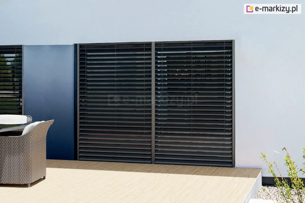 Nowoczesna fasada domu z dwoma dużymi przeszkleniami wyposażonymi w żaluzje fasadowe zewnętrzne w kolorze antracytowym - Z90 BOX podtynkowy. System żaluzji z poziomymi lamelami aluminiowymi, osadzony we wnękach okiennych w elewacji w jasnym kolorze. Po lewej stronie widoczne krzesło technorattanowe z białą poduszką na drewnianym tarasie. Część elewacji w ciemnoniebieskim wykończeniu kontrastuje z bielą tynku. Przed oknami widać precyzyjnie zamontowane żaluzje, których lamele są ustawione w pozycji półotwartej, umożliwiając kontrolę dopływu światła słonecznego.