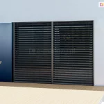 Nowoczesna fasada domu z dwoma dużymi przeszkleniami wyposażonymi w żaluzje fasadowe zewnętrzne w kolorze antracytowym - Z90 BOX podtynkowy. System żaluzji z poziomymi lamelami aluminiowymi, osadzony we wnękach okiennych w elewacji w jasnym kolorze. Po lewej stronie widoczne krzesło technorattanowe z białą poduszką na drewnianym tarasie. Część elewacji w ciemnoniebieskim wykończeniu kontrastuje z bielą tynku. Przed oknami widać precyzyjnie zamontowane żaluzje, których lamele są ustawione w pozycji półotwartej, umożliwiając kontrolę dopływu światła słonecznego.