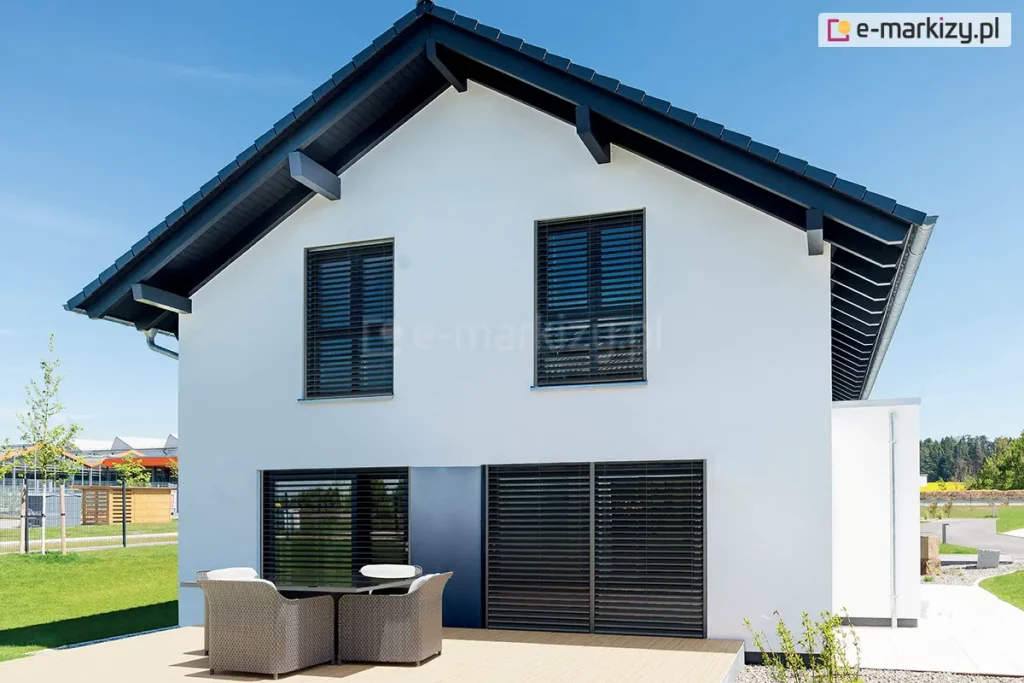 Żaluzje fasadowe Z90 Box w kolorze czarnym, zamontowane w otworach drzwi i okien piętrowego budynku o białej fasadzie, antracytowej stolarce okiennej oraz czarnym dachu. Przed budynkiem widoczny stół z czterema dużymi fotelami ratanowymi na niewielkim tarasie. W tle ogród i sąsiednie budynki. Nad domkiem jasne niebieskie niebo.