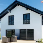 Żaluzje fasadowe Z90 Box w kolorze czarnym, zamontowane w otworach drzwi i okien piętrowego budynku o białej fasadzie, antracytowej stolarce okiennej oraz czarnym dachu. Przed budynkiem widoczny stół z czterema dużymi fotelami ratanowymi na niewielkim tarasie. W tle ogród i sąsiednie budynki. Nad domkiem jasne niebieskie niebo.