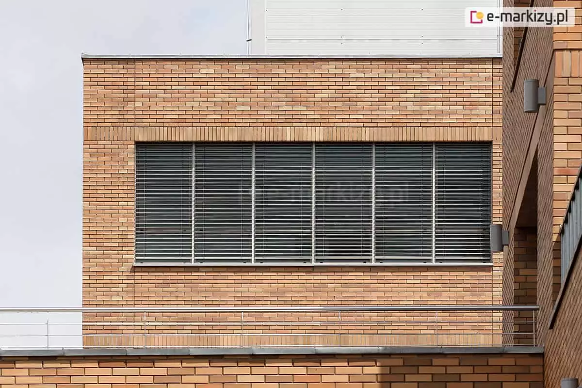 Elewacja nowoczesnego budynku z cegły klinkierowej w kolorze piaskowym, z rzędem dużych okien wyposażonych w zewnętrzne żaluzje fasadowe z poziomymi, aluminiowymi lamelami. Żaluzje zamontowane są na całej długości przeszklenia, z podziałem na segmenty okienne oddzielone pionowymi profilami. Lamele ustawione są w pozycji półotwartej, co pozwala na kontrolę dostępu światła do wnętrza. W dolnej części zdjęcia widoczny jest stalowy balkon z balustradą, a po prawej stronie fragment wnęki z lampami elewacyjnymi. Styl budynku sugeruje zastosowanie w obiektach biurowych lub użyteczności publicznej.