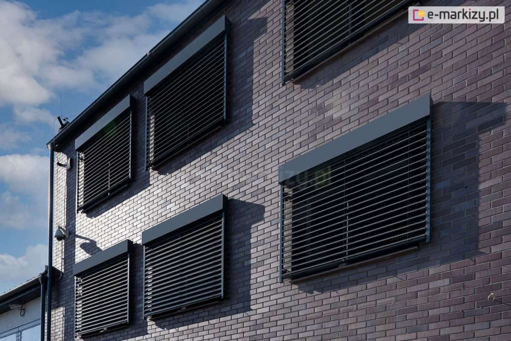 Widok na boczno-frontową elewację nowoczesnego budynku wielorodzinnego lub biurowego, wyłożoną ciemną cegłą klinkierową. Na ścianie zamontowanych jest sześć identycznych żaluzji fasadowych Z90 Cube w kolorze antracytowym, rozmieszczonych symetrycznie w dwóch rzędach. Wszystkie żaluzje są opuszczone, a ich lamele ustawione w pozycji poziomej, co tworzy powtarzalny, geometryczny wzór na fasadzie. Budynek posiada ciemne rynny i obróbki blacharskie. W tle widoczne jest pogodne, błękitne niebo z lekkim zachmurzeniem. Zdjęcie prezentuje estetykę nowoczesnych systemów osłonowych zintegrowanych z architekturą obiektu.