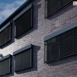 Widok na boczno-frontową elewację nowoczesnego budynku wielorodzinnego lub biurowego, wyłożoną ciemną cegłą klinkierową. Na ścianie zamontowanych jest sześć identycznych żaluzji fasadowych Z90 Cube w kolorze antracytowym, rozmieszczonych symetrycznie w dwóch rzędach. Wszystkie żaluzje są opuszczone, a ich lamele ustawione w pozycji poziomej, co tworzy powtarzalny, geometryczny wzór na fasadzie. Budynek posiada ciemne rynny i obróbki blacharskie. W tle widoczne jest pogodne, błękitne niebo z lekkim zachmurzeniem. Zdjęcie prezentuje estetykę nowoczesnych systemów osłonowych zintegrowanych z architekturą obiektu.