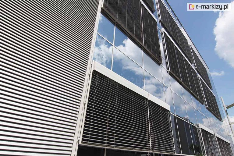 Perspektywiczne ujęcie nowoczesnej fasady budynku biurowego o dużej powierzchni przeszkleń, na której zamontowano system zewnętrznych żaluzji fasadowych C80 OVAL. Lewa część zdjęcia przedstawia gęsto rozmieszczone, poziome lamele w kolorze srebrnym, całkowicie zasłaniające elewację. Środkowa i prawa część obrazu ukazuje duże tafle szkła, w których odbija się błękitne niebo z białymi chmurami. Na poszczególnych modułach okiennych widoczne są opuszczone żaluzje fasadowe w ciemniejszym kolorze, zamontowane w dedykowanych kasetach osłonowych o charakterystycznym zaokrąglonym kształcie. Systemy te są wyposażone w prowadnice boczne utrzymujące stabilność konstrukcji na wietrze. Ujęcie wykonane z dołu ku górze podkreśla rytmiczność i geometrię nowoczesnej architektury.