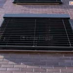 Zewnętrzna żaluzja fasadowa Z90 Cube w kolorze ciemnego antracytu zamontowana na elewacji wykonanej z klinkierowych cegieł w odcieniach brązu i fioletu. Żaluzja jest opuszczona i zasłania okno, a jej lamele są ustawione poziomo, pozwalając na częściowy dopływ światła do wnętrza. Konstrukcja zawiera prowadnice boczne oraz górną kasetę osłonową dopasowaną kolorystycznie do lameli. Poniżej dolnej krawędzi żaluzji widoczny jest zewnętrzny parapet. Zdjęcie wykonane pod kątem z dołu, ukazujące detal montażu systemu osłonowego na ceglanej ścianie budynku.